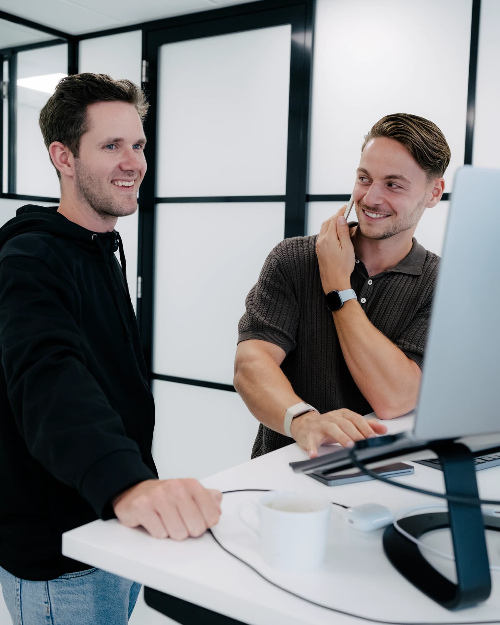 Iwan en Sam staan bij Sams werkplek terwijl Sam een klant aan de telefoon heeft